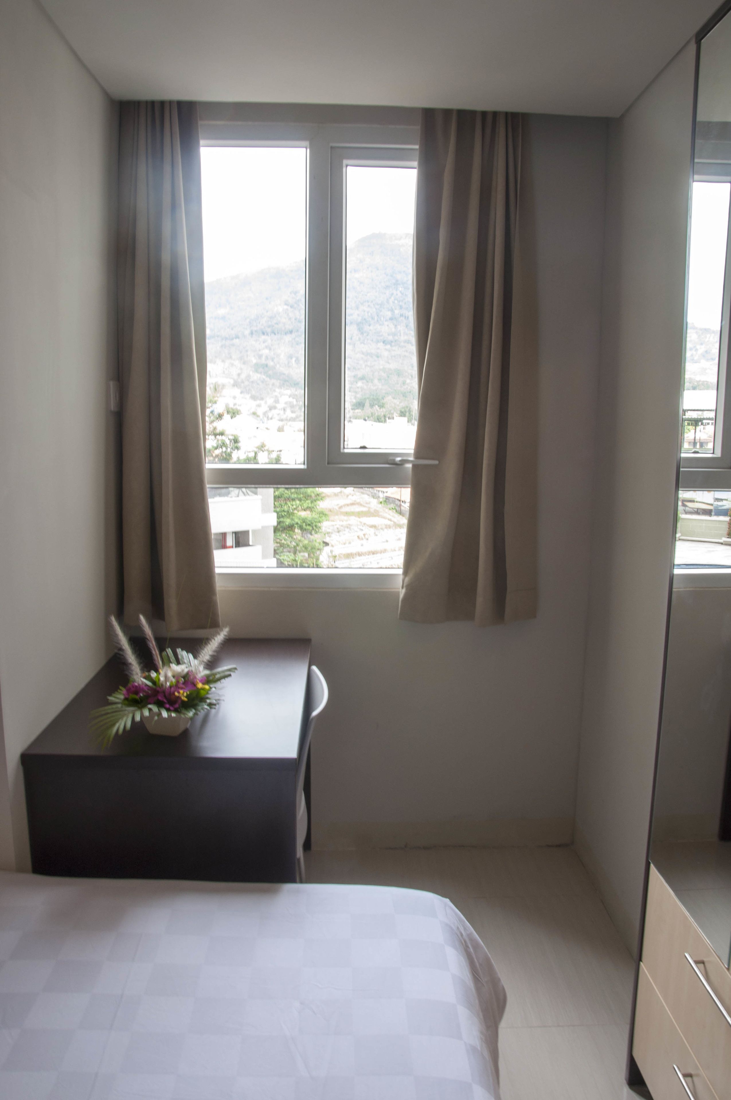 A hotel room with a bed, a small table with a vase of flowers, a window with curtains, and a dresser.
