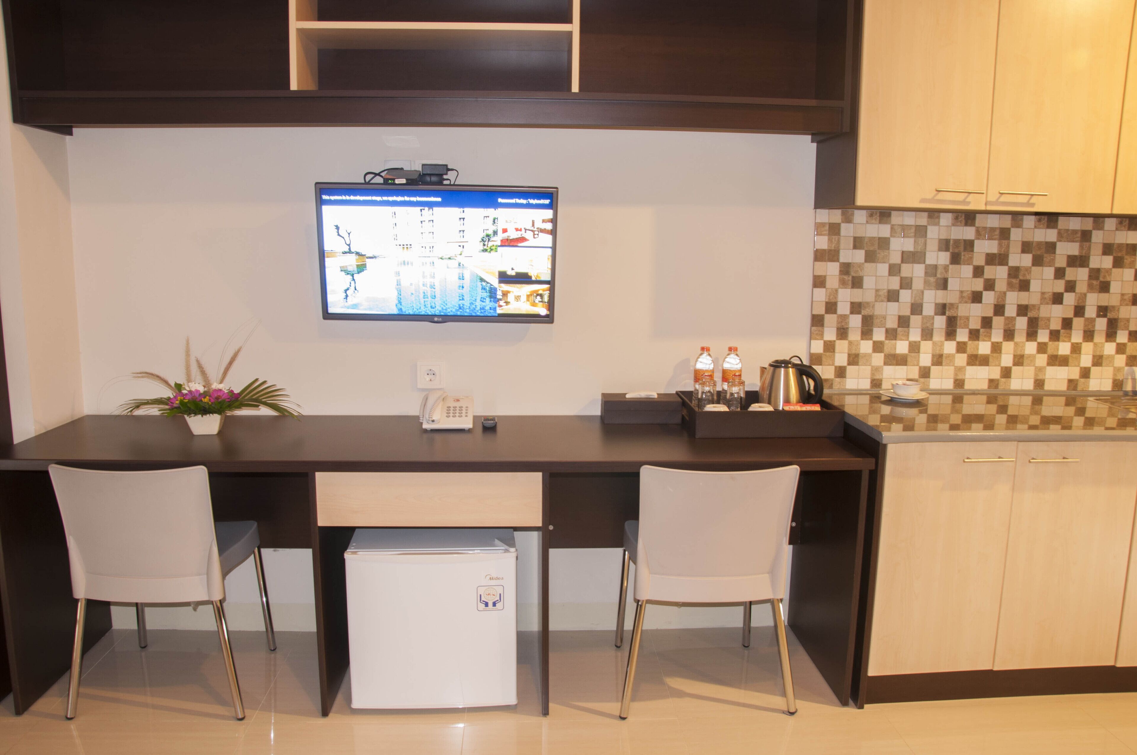 Una cocina compacta con refrigerador incorporado, dos sillas blancas, una mesita y un televisor de pantalla plana montado en la pared.