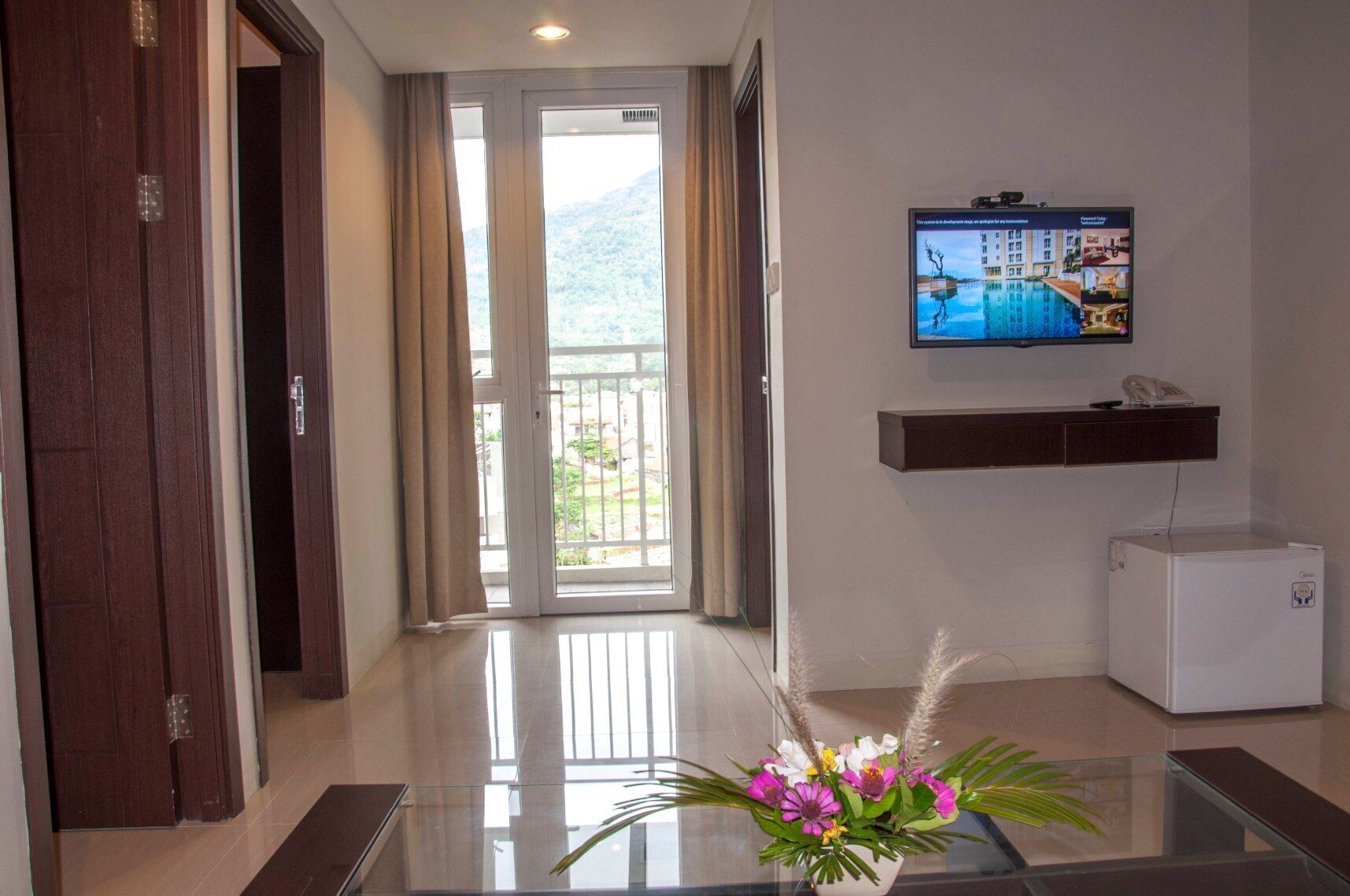A hotel room with a glass table, a TV on the wall, a mini-fridge, and a view of the outdoors.