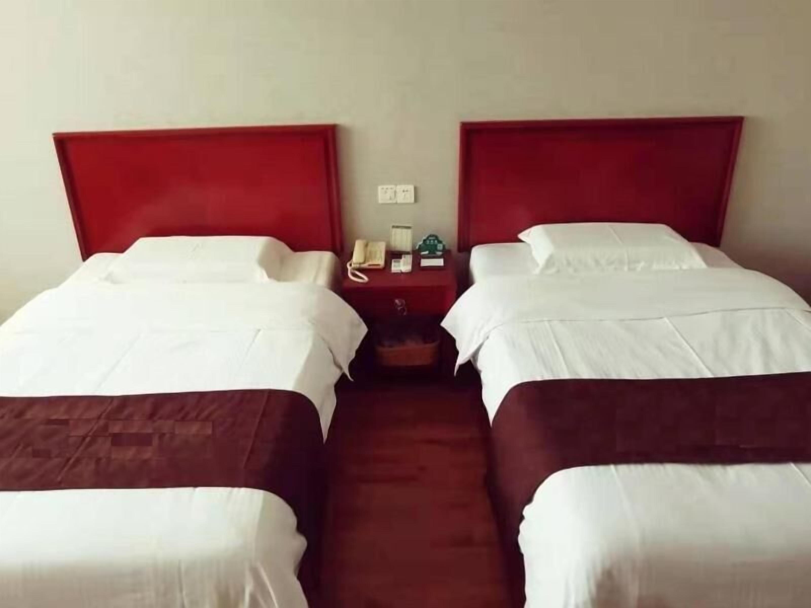 A hotel room with two single beds, each with a red headboard, white bedding, and a small bedside table with a telephone and a green bottle.