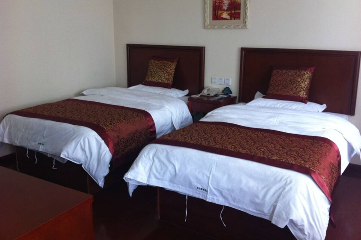 Ein Hotelzimmer mit zwei Einzelbetten, jeweils mit weißer Bettdecke und einer Decke in Rot und Gold. Es gibt ein hölzernes Kopfteil, einen Nachttisch mit Telefon und ein gerahmtes Bild an der Wand.