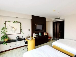Habitación de hotel con dos camas, un escritorio con computadora, un televisor montado en la pared y una planta pequeña.