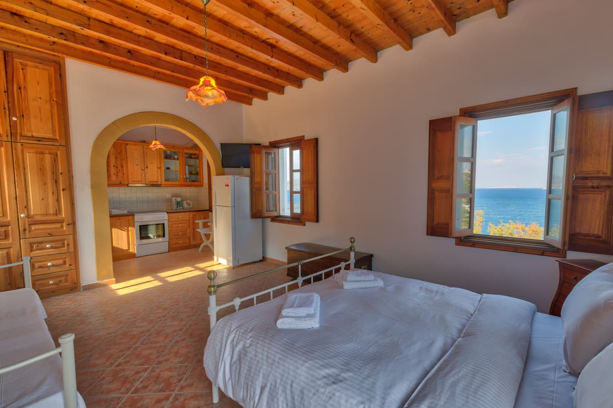 A bedroom with a large bed, a view of the sea through a window, wooden ceiling beams, and a kitchen in the background.