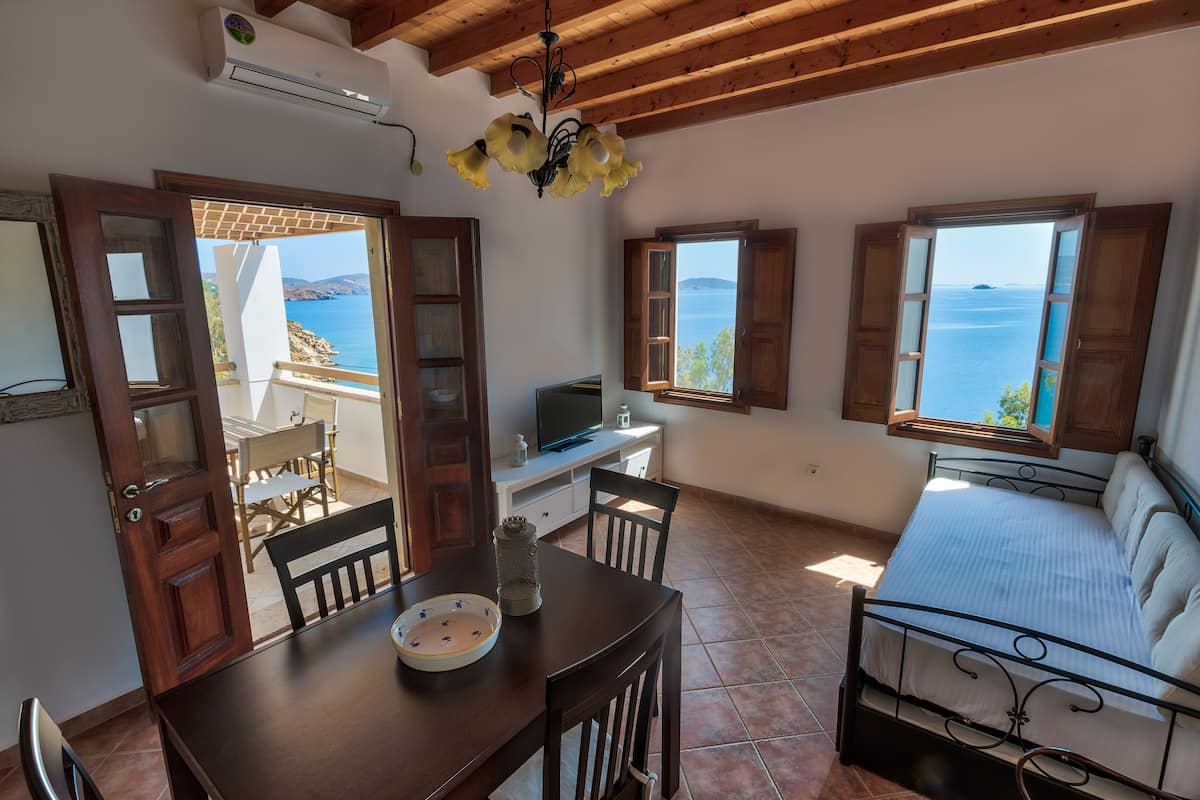 A room with a bed, a dining table with chairs, a TV, and a view of the sea.