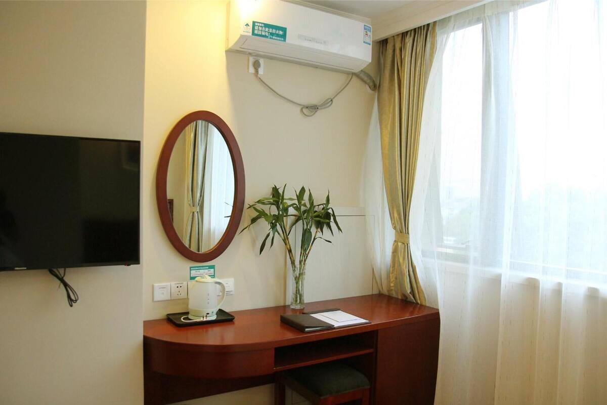A hotel room with a flat-screen TV mounted on the wall, a wooden desk with a mirror, a small plant, and a window with sheer curtains.