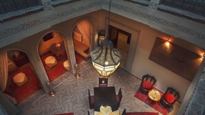 An interior view of a hotel with a dining area featuring a table with red cushions, a hanging lantern, and a view of a room with a bed and a red carpet. - Riad La Kahana (Marrakech)