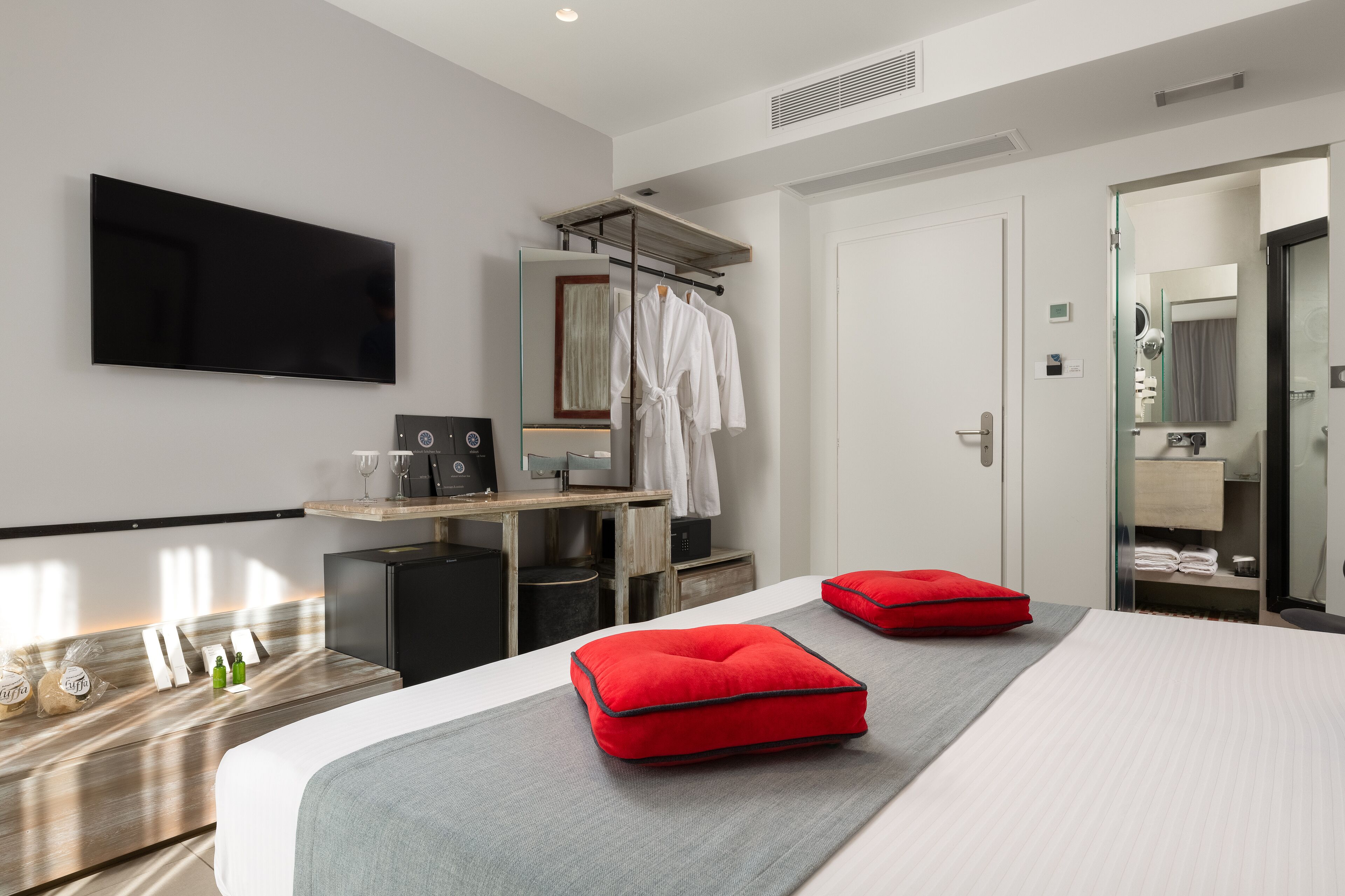A modern hotel room with a large bed, a flat-screen TV mounted on the wall, a desk with a computer, and a mirror.