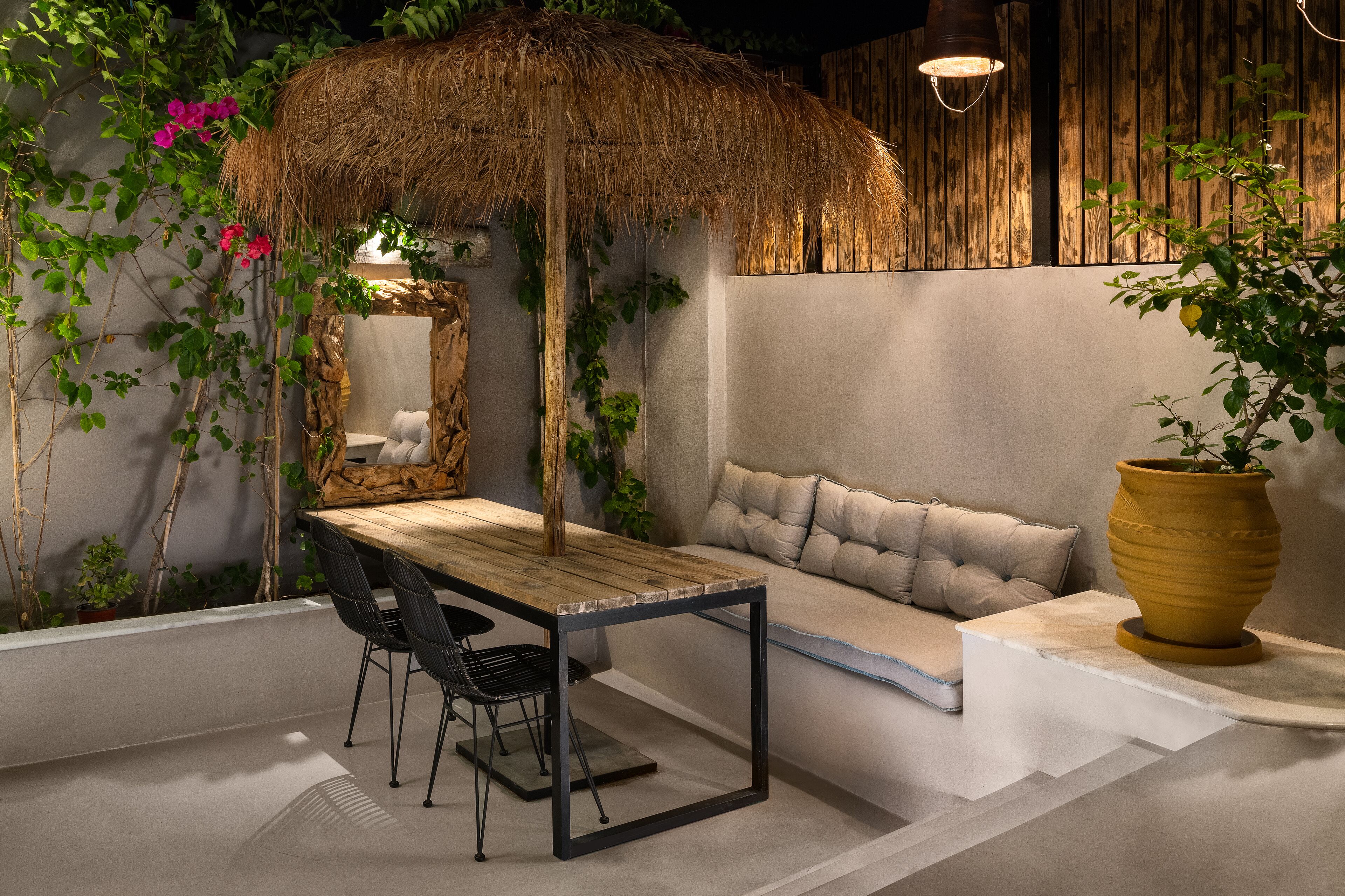 A cozy outdoor seating area with a wooden table and chairs, a cushioned bench, and a large potted plant.