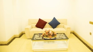 Una habitación con un sofá beige, dos almohadas, una mesa de centro de vidrio y un arreglo floral, además de un piso de baldosas.