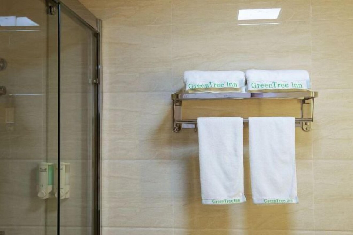 Un baño con puerta de ducha de cristal, dos toallas blancas colgadas en un perchero y un dispensador de jabón en la pared.