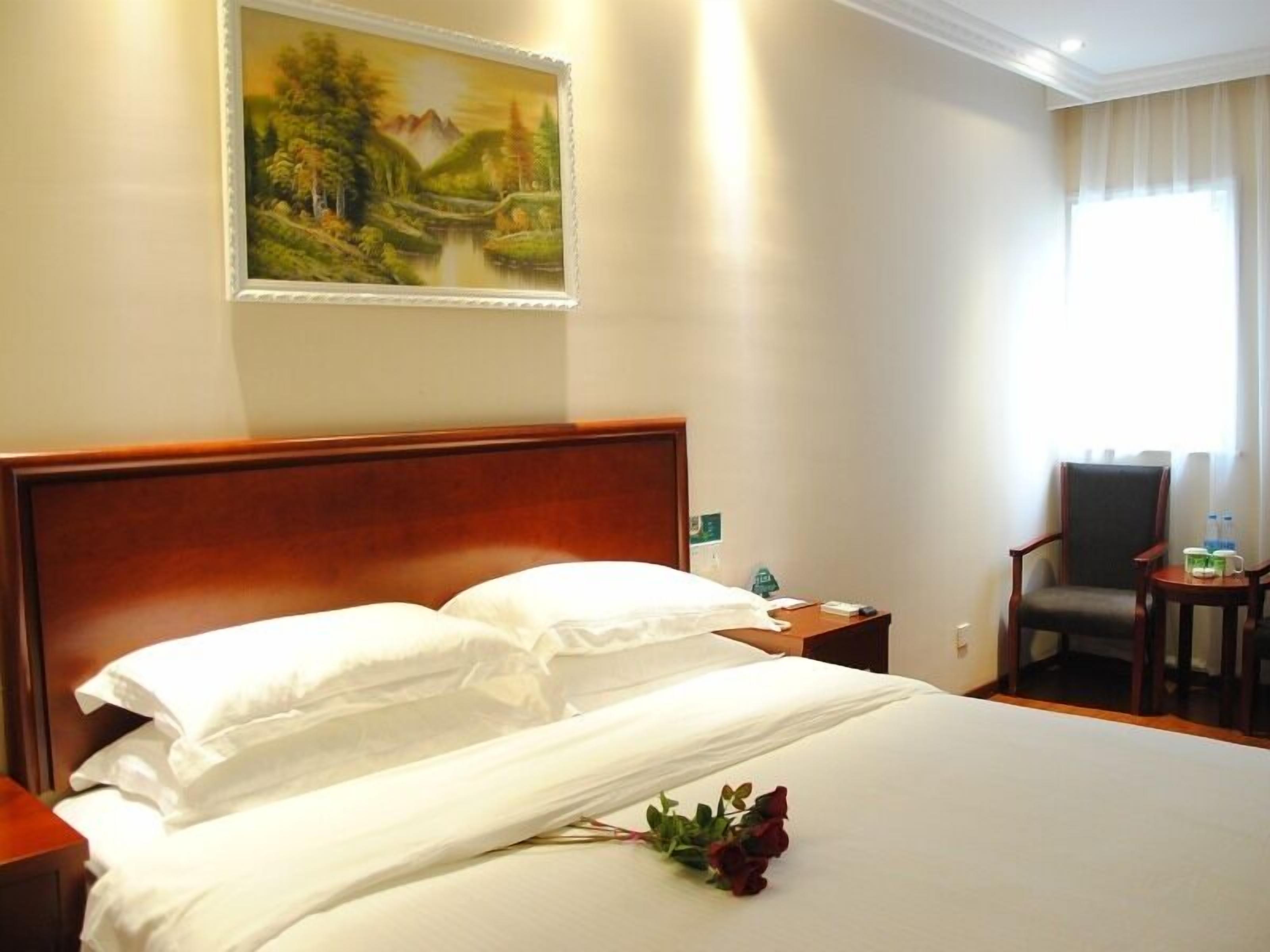 A hotel room with a large bed, a wooden headboard, a painting of a landscape on the wall, a chair, and a small table with a bottle and a glass.