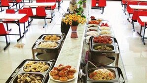 Montaje de bufé con diversas opciones de comida, como pasta, ensaladas y otros platos, dispuesto en una mesa larga en un comedor con sillas rojas.