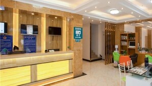 Un espace d’accueil comprenant un comptoir de réception en bois, un téléviseur fixé au mur, un panneau de signalisation vert et un coin salon avec des chaises et une table.