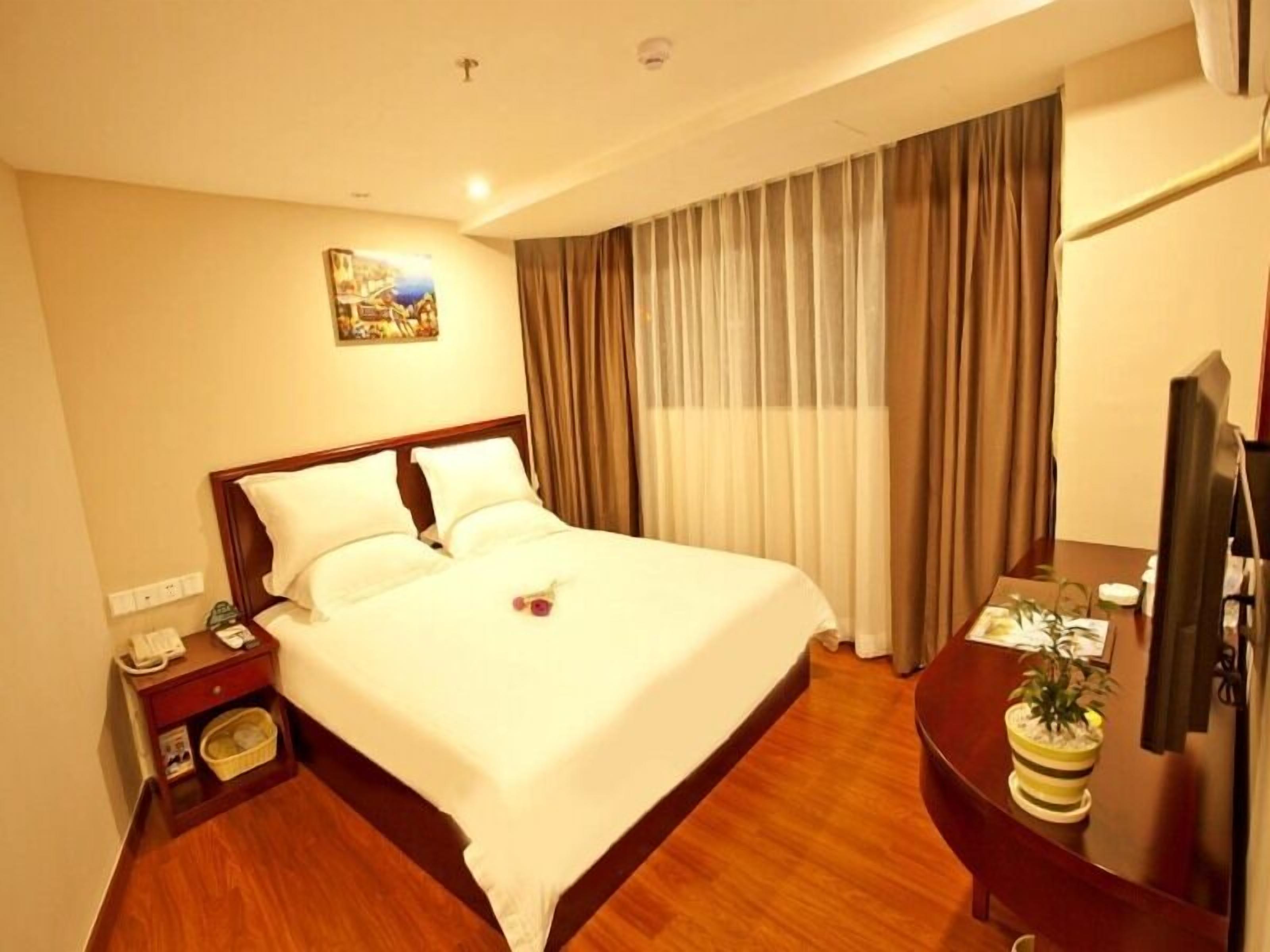 A hotel room with a large bed, a bedside table with a lamp, a desk with a computer, and a potted plant.