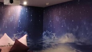 Ein Schlafzimmer mit Wandgemälde eines Sternenhimmels, einem Bett mit weißer Bettwäsche und einem Kopfkissen mit Aufschrift.