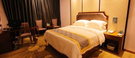 A hotel room with a large bed, a desk with a computer, two chairs, and a small trash bin.