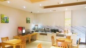 A dining area with wooden tables and chairs, a kitchen in the background, and a ceiling with recessed lighting.