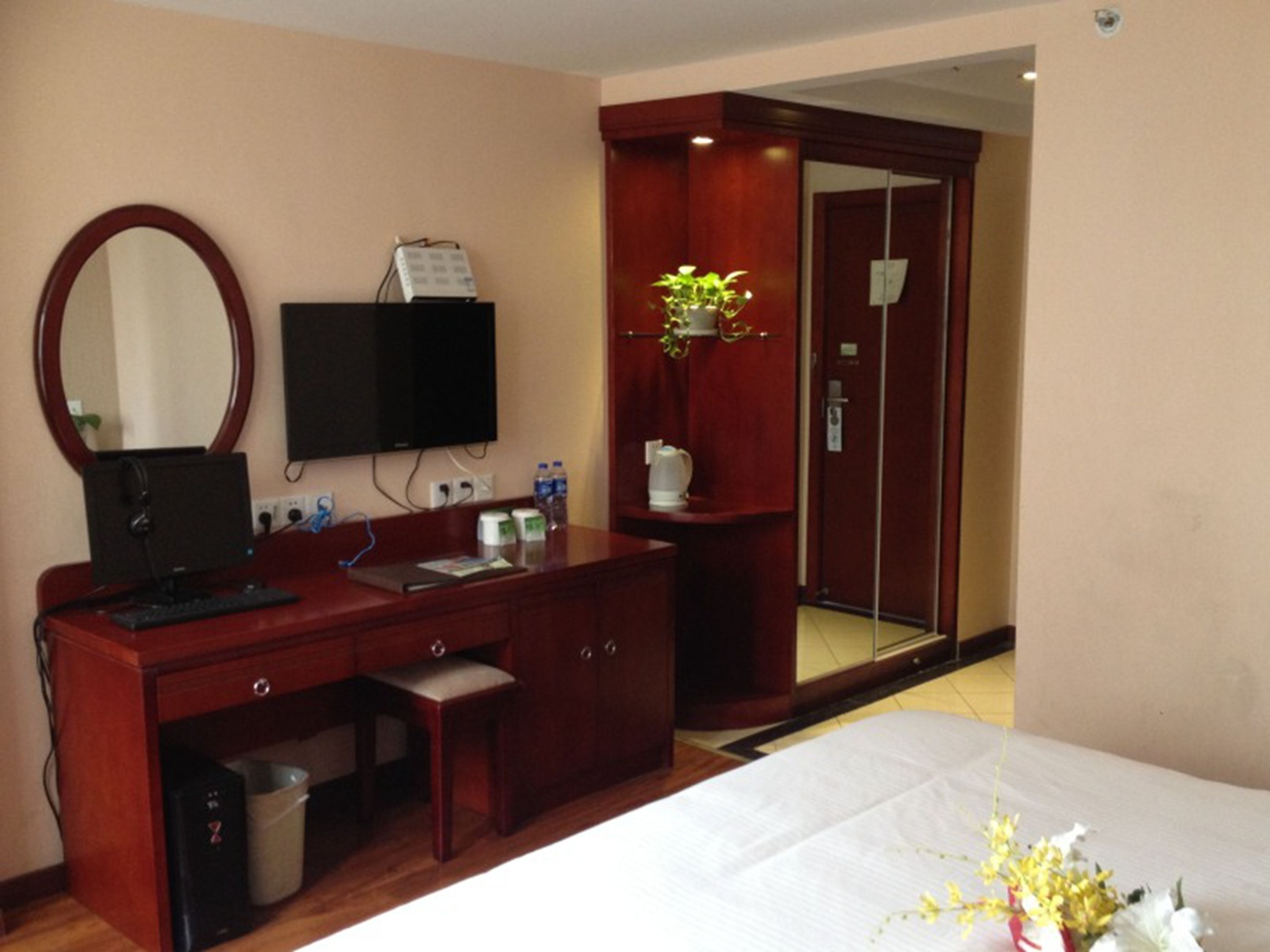 A hotel room with a wooden desk, a computer, a television, a bed, a mirror, a small trash bin, a water dispenser, and a plant.