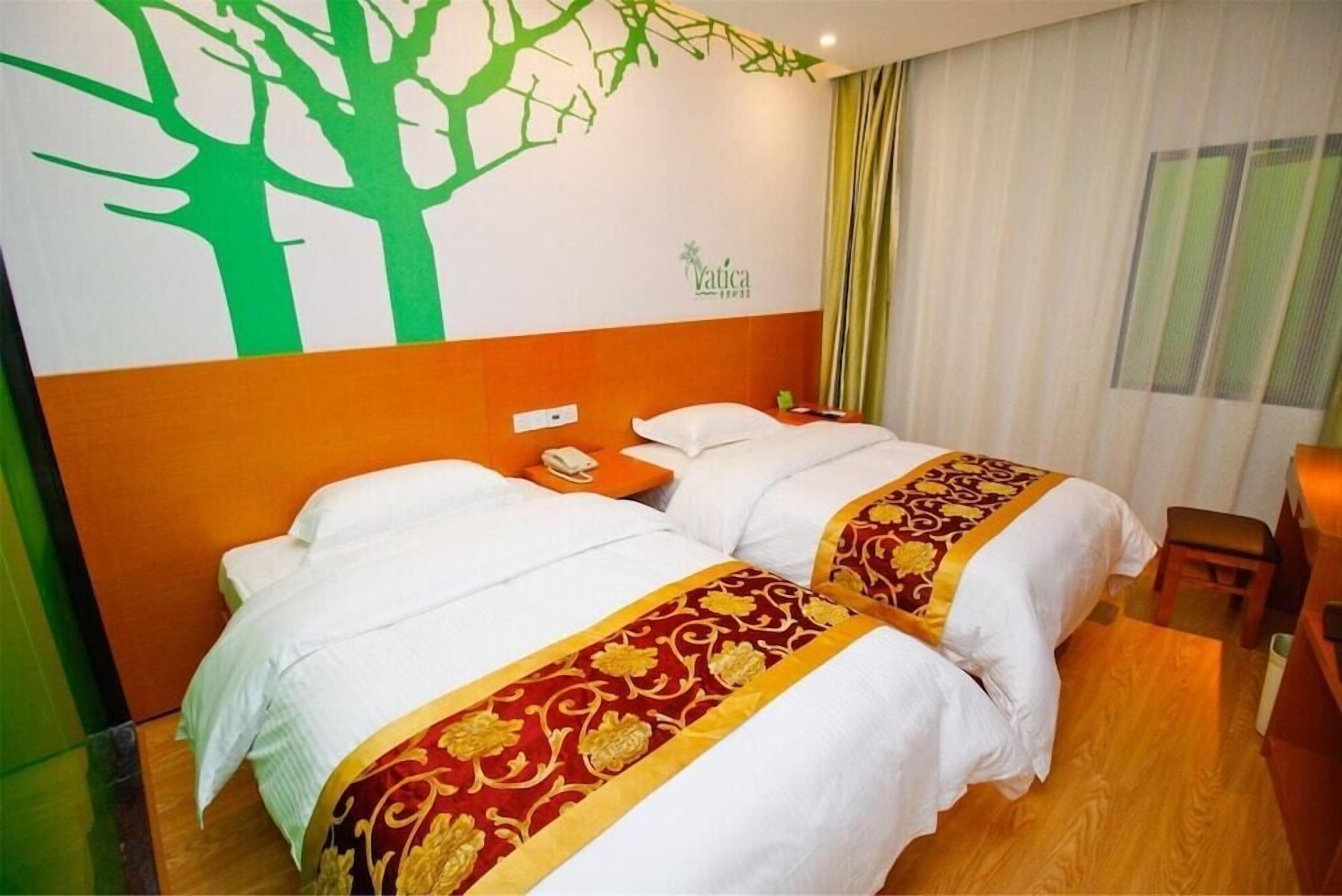 A hotel room with two beds, each with a white and red bedspread, a wall mural of a tree, and a window with curtains.