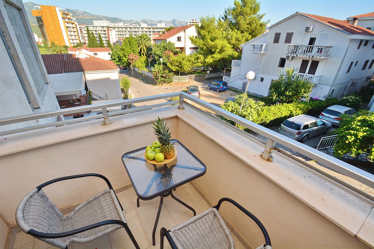 A balcony with two wicker chairs, a small table with fruit, and a view of a residential area with buildings and parked cars.