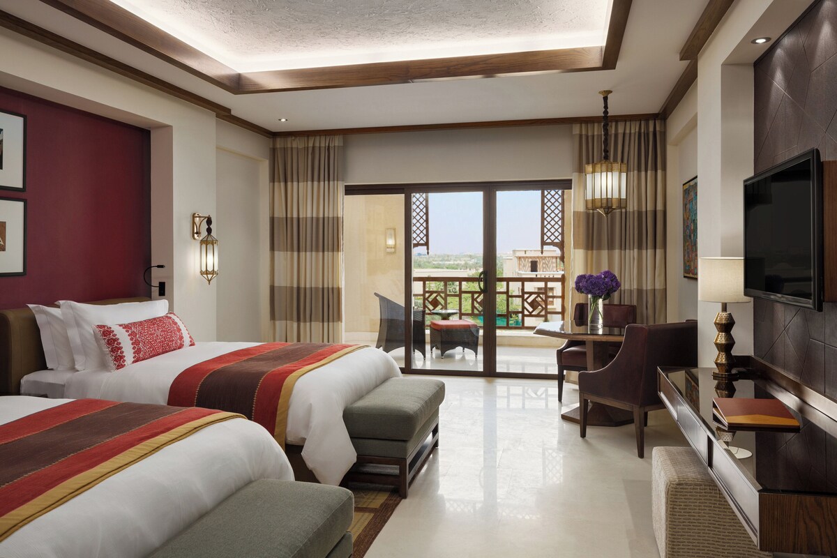 A hotel room with two beds, a desk, a chair, a TV, and a balcony with a view.