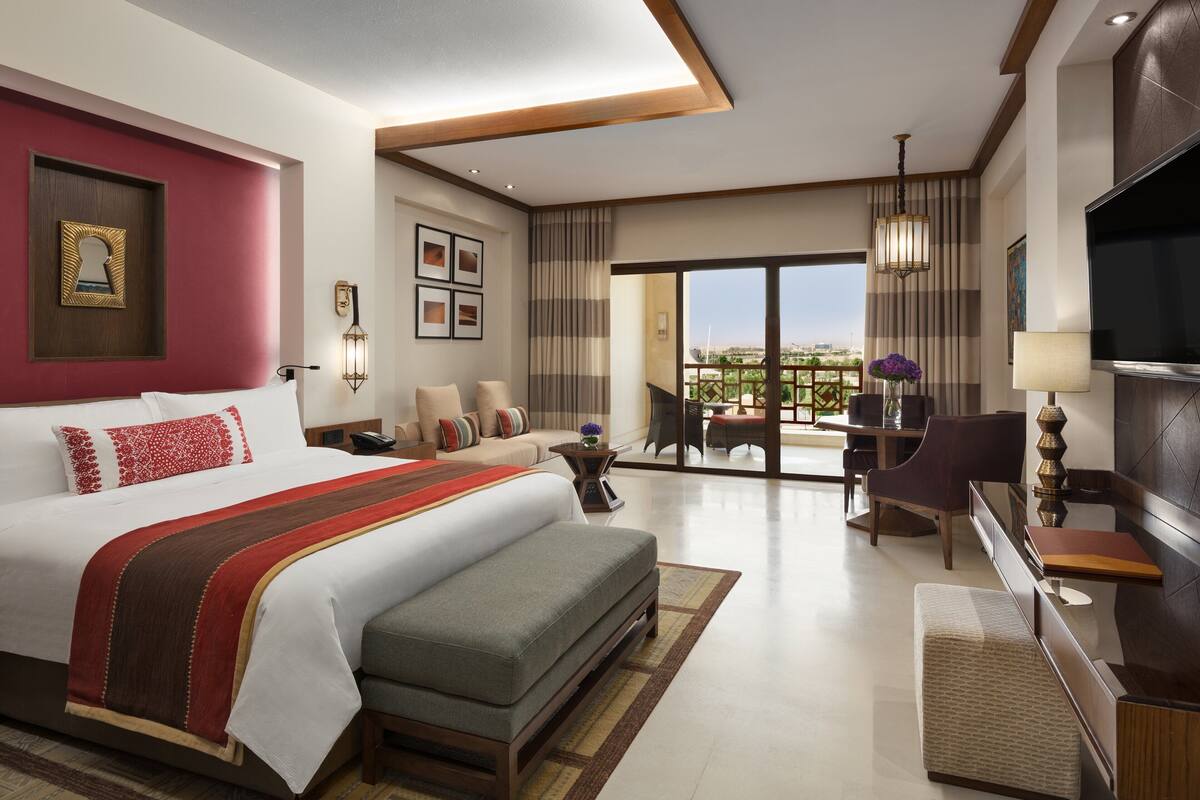 A modern hotel room with a large bed, a seating area, a TV, and a balcony with a view.