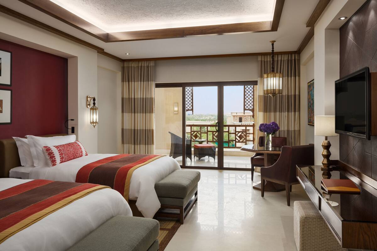 A hotel room with two beds, a desk, a chair, a TV, and a balcony with a view.