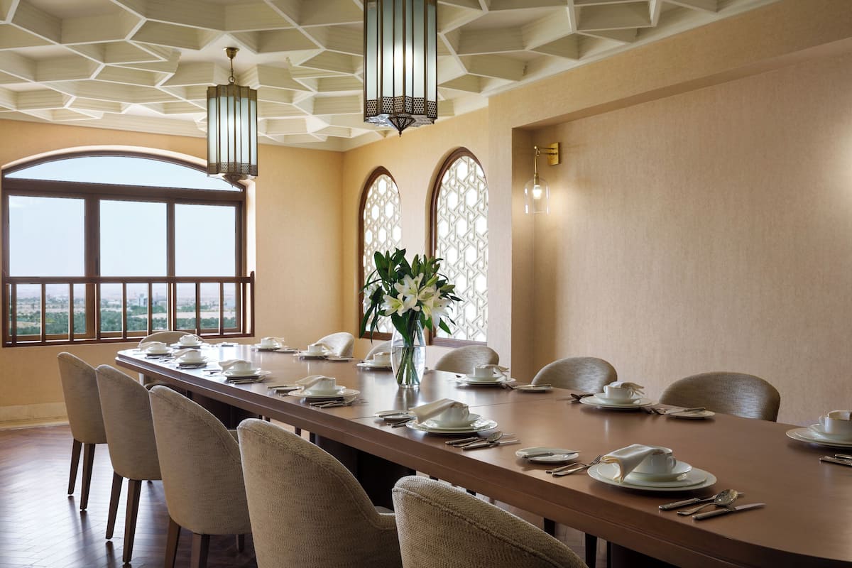 A long dining table set for a meal with chairs, a vase with flowers, and a view through large windows.
