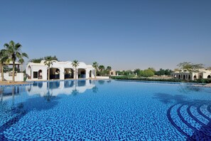 A large, clear blue pool with a tiled pattern, surrounded by white buildings with arches and palm trees.