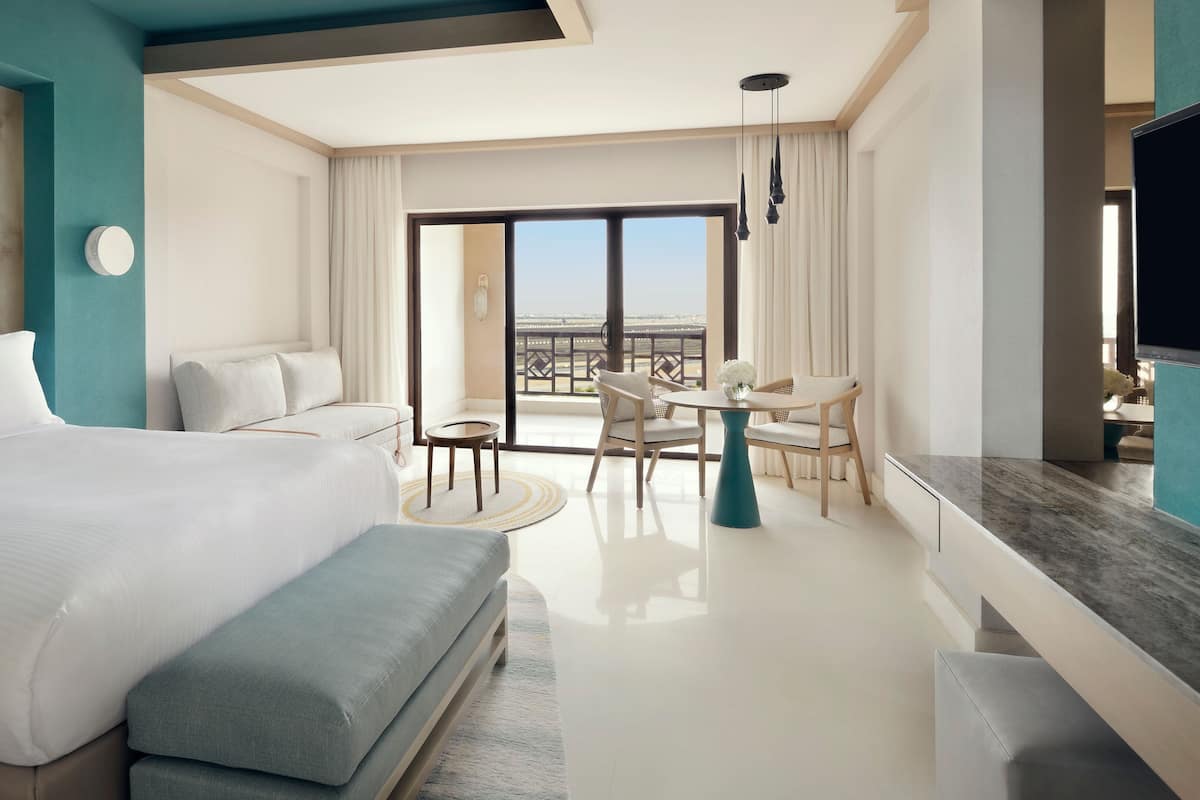 A modern hotel room with a large bed, a seating area with a table, and a balcony with a view.