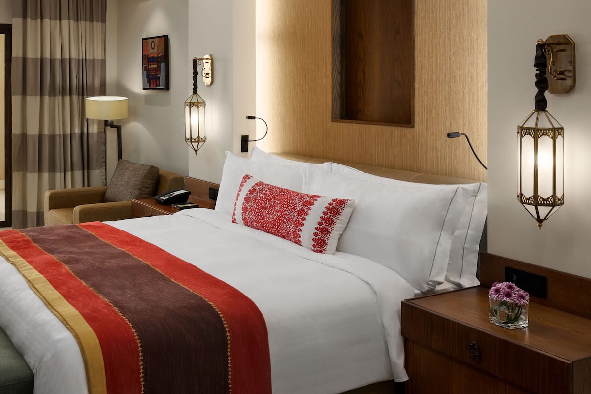 A hotel room with a large bed, a wooden headboard, a nightstand with a lamp, and a small table with a vase of flowers.
