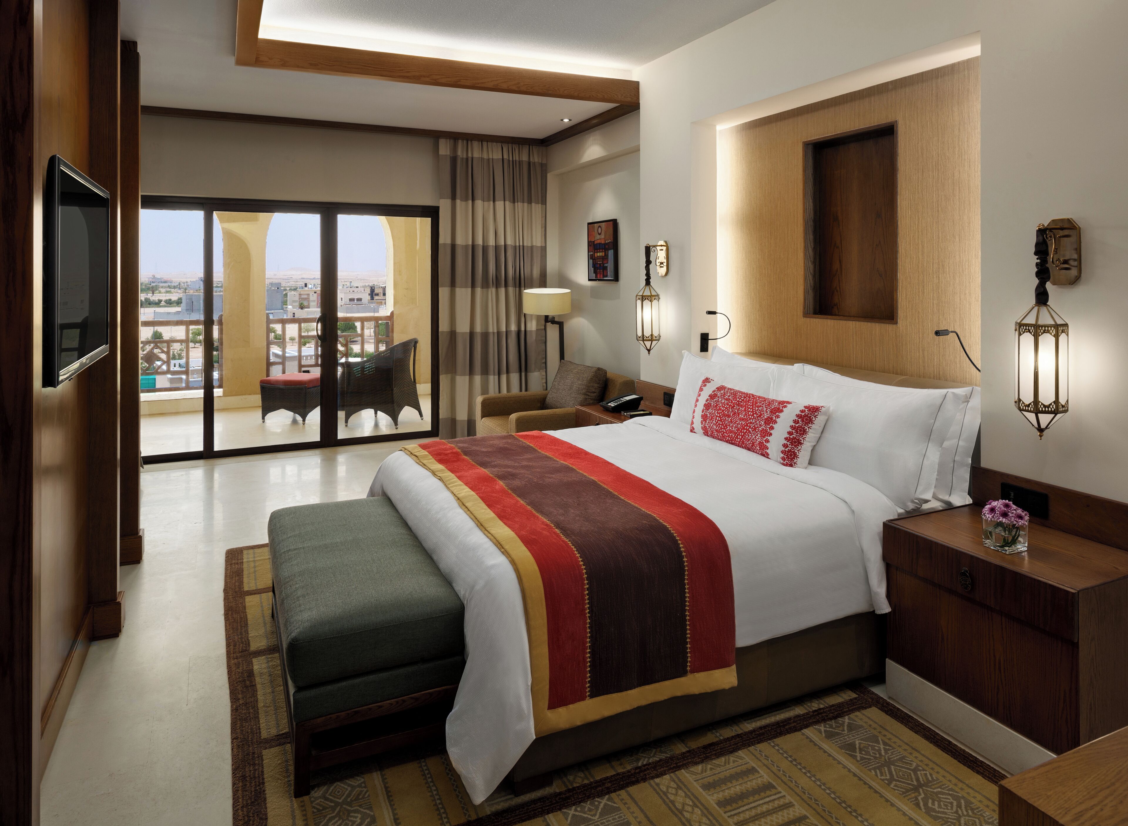 A hotel room with a large bed, a flat-screen TV, a balcony with a view, and a wooden headboard.