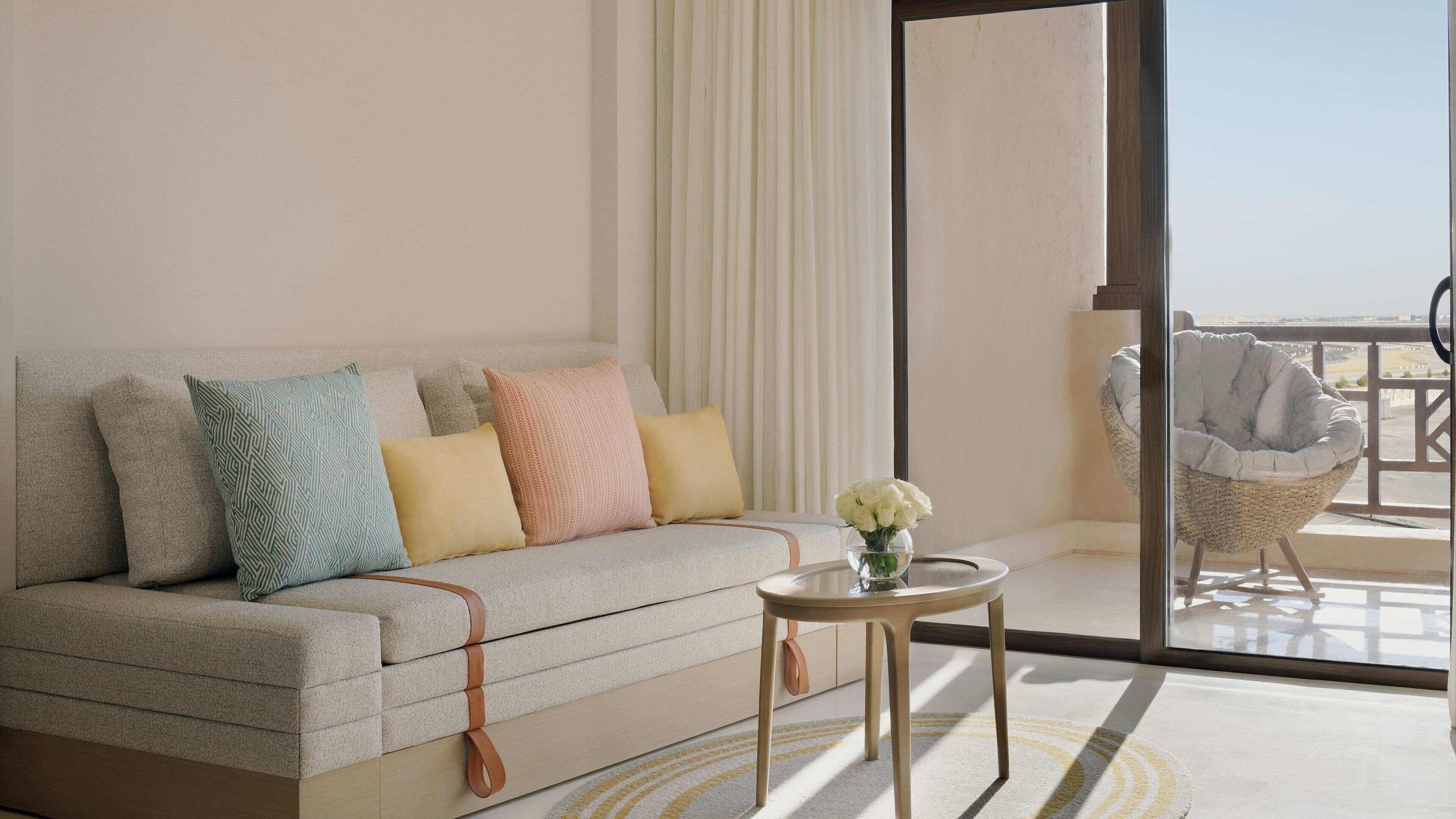 A modern living room with a sofa, pillows, a small table with a vase, and a view of the outdoors.