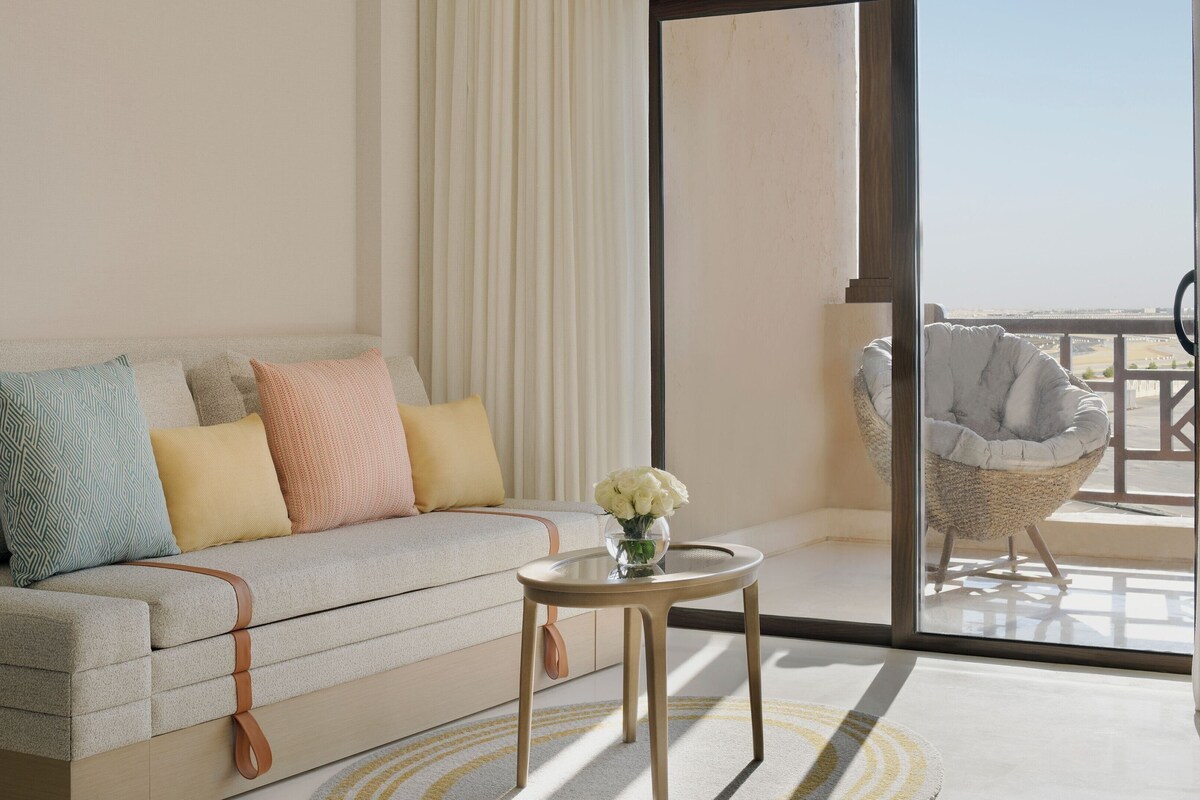 A modern living room with a sofa, pillows, a small table with a vase, and a view of the outdoors.