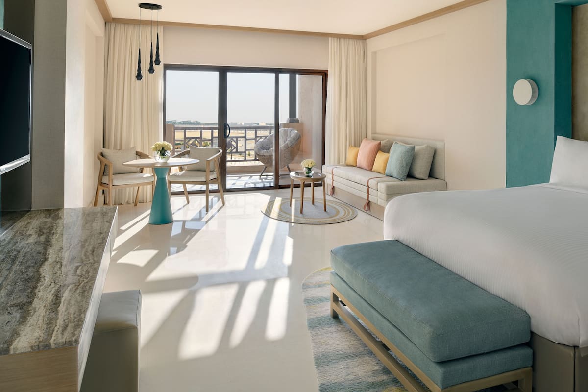 A modern hotel room with a large bed, a sofa, a dining table, and a balcony with a view.