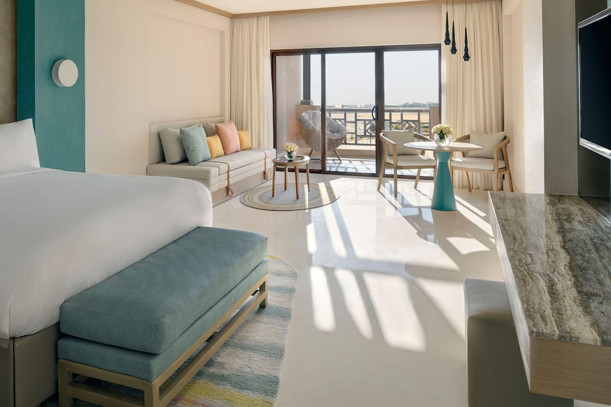 A hotel room with a large bed, a sofa, a dining area with a table and chairs, and a balcony with a view.