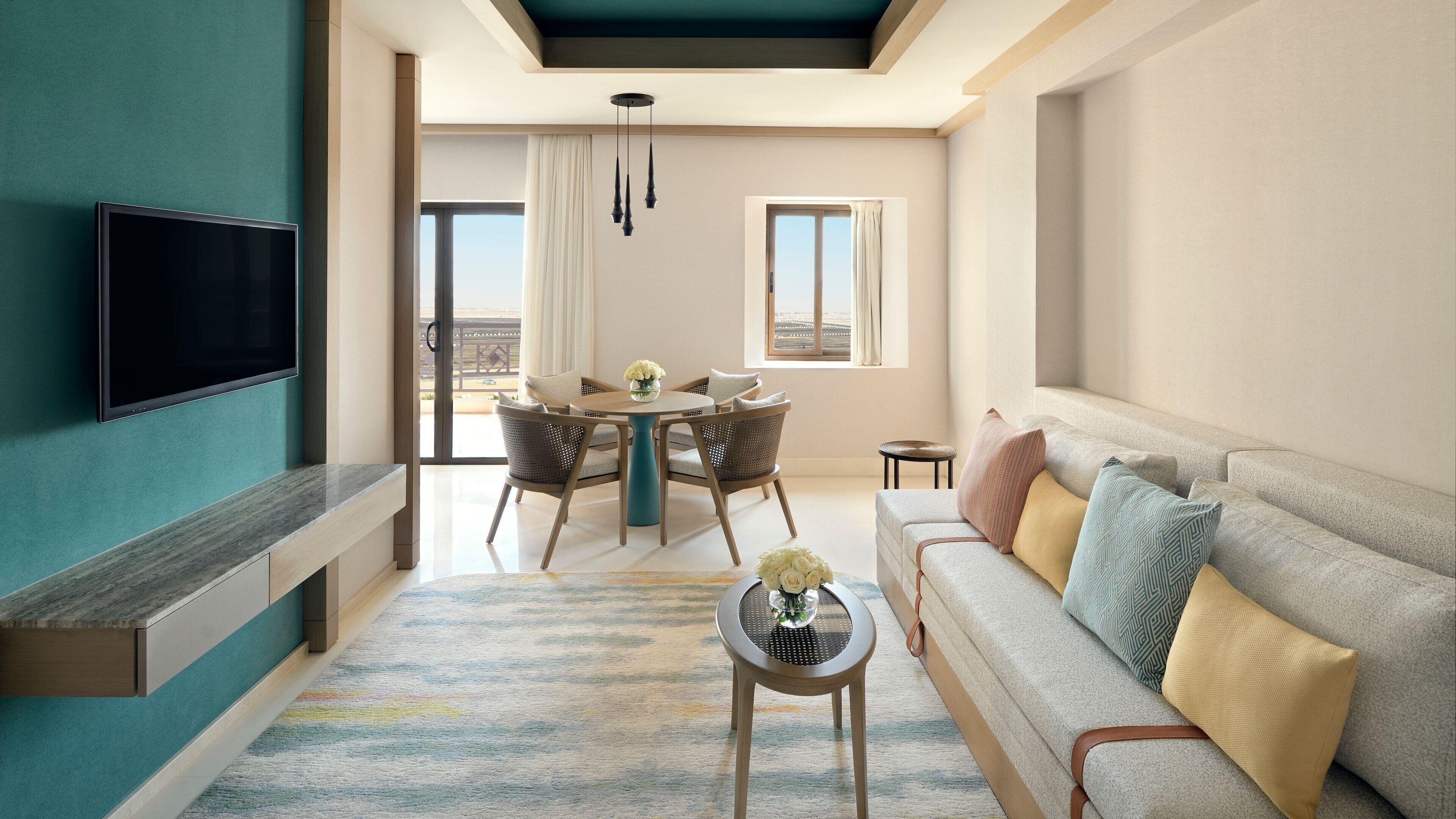 A modern living room with a flat-screen TV, a sofa with patterned cushions, a dining area with wicker chairs, and a view of the outdoors through large windows.