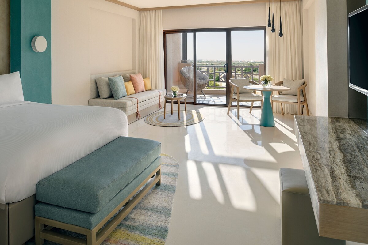 A modern hotel room with a large bed, a sofa, a dining area, and a balcony with a view.