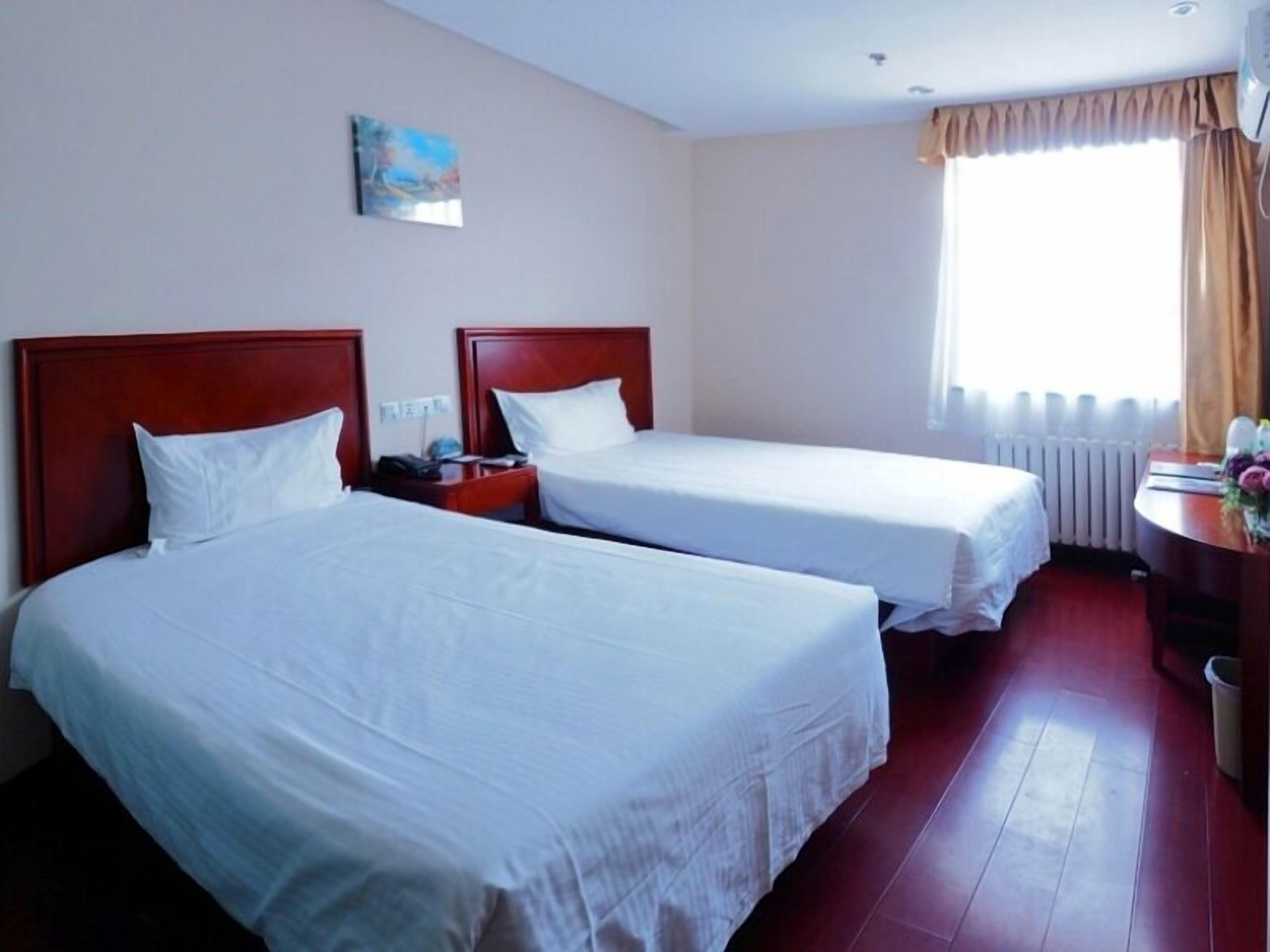 A hotel room with two single beds, a wooden headboard, a small bedside table, a radiator, and a window with curtains.