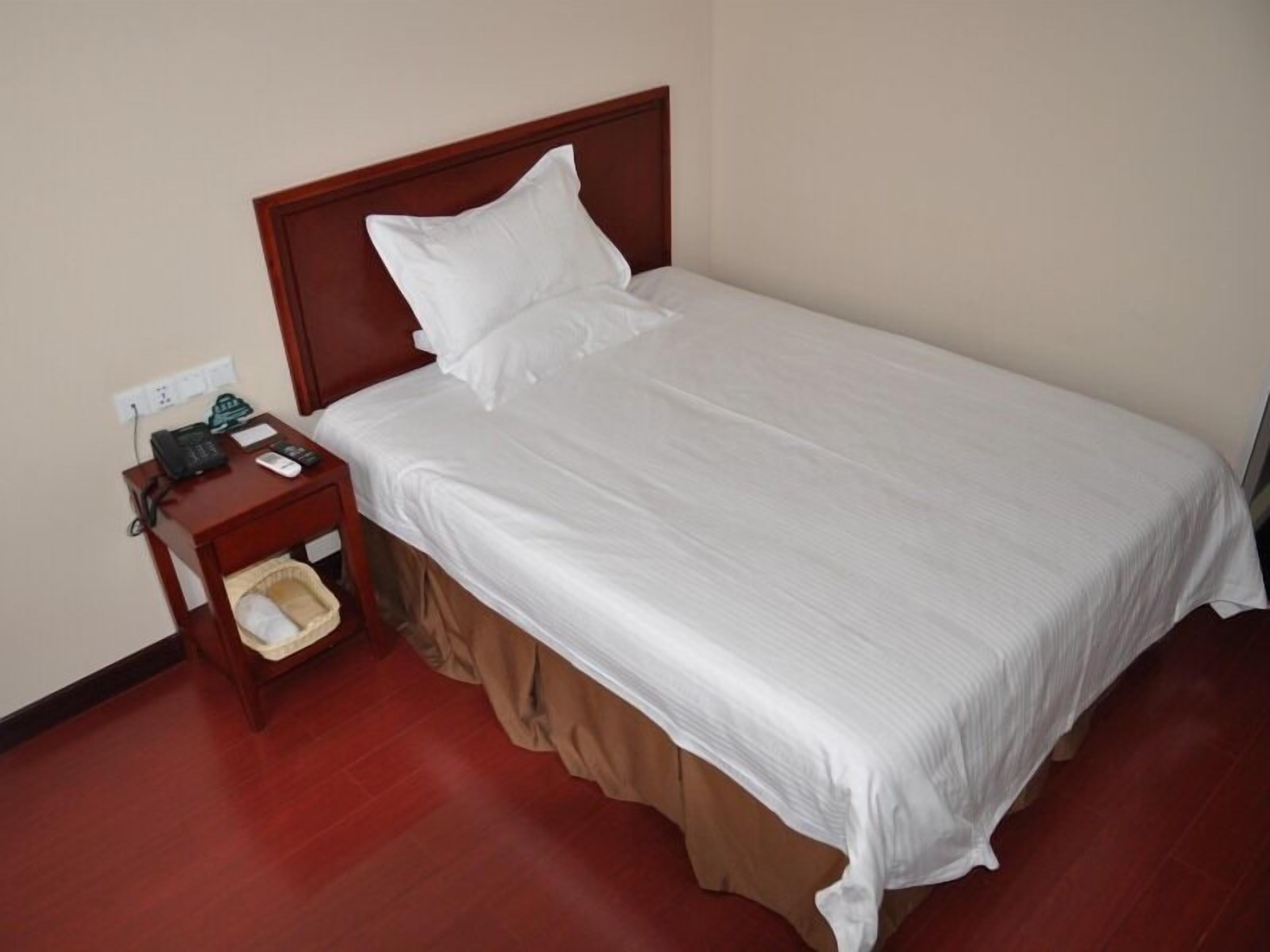 A neatly made bed with white bedding, a wooden nightstand with a telephone and a trash bin, and a brown skirt on the bed.