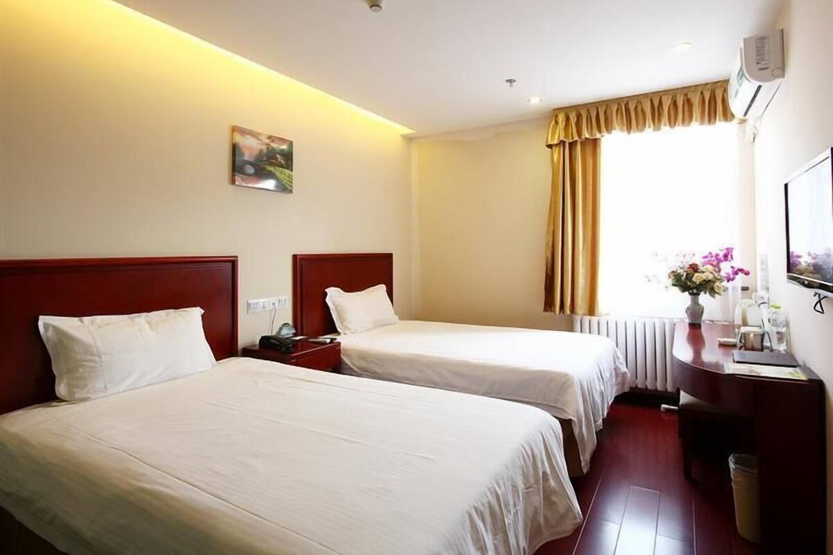 Une chambre d’hôtel avec deux lits simples, un bureau avec une chaise, un radiateur, une fenêtre avec des rideaux et un climatiseur fixé au mur.