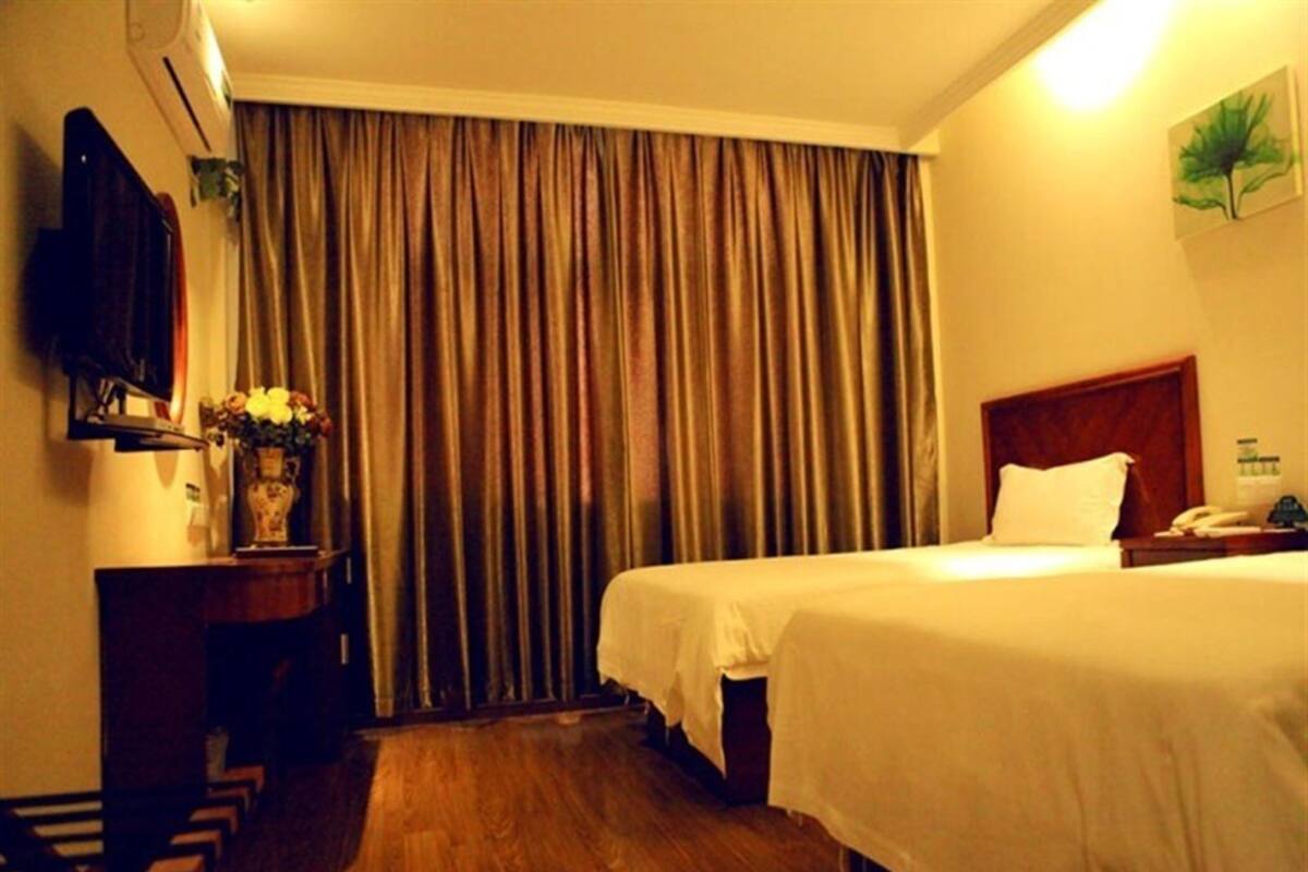 A hotel room with two beds, a wooden floor, a large window with curtains, a flat-screen TV, and a small table with a vase of flowers.