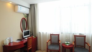 A hotel room with a desk, two chairs, a small table with drinks, a TV, and an air conditioning unit.