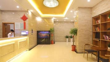 A hotel lobby with a reception desk, a television, potted plants, and a bookshelf.