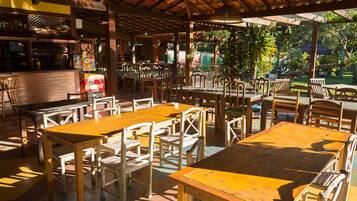 An outdoor dining area with wooden tables and chairs, a brick counter, and a view of greenery.