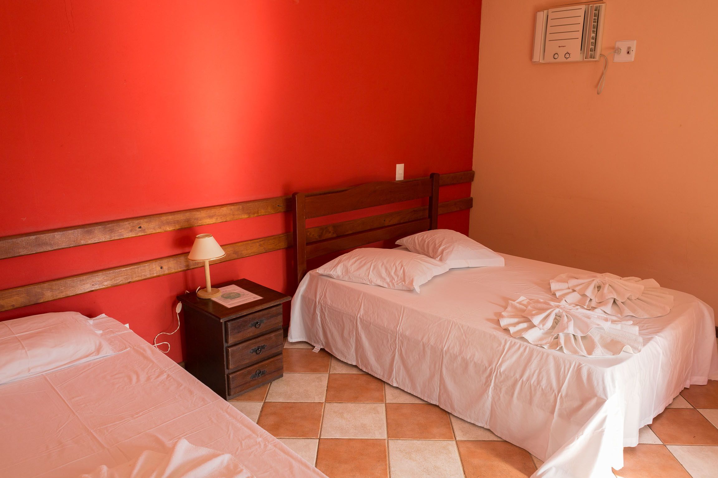A bedroom with two beds, a wooden headboard, a nightstand with a lamp, and a wall-mounted electrical outlet.