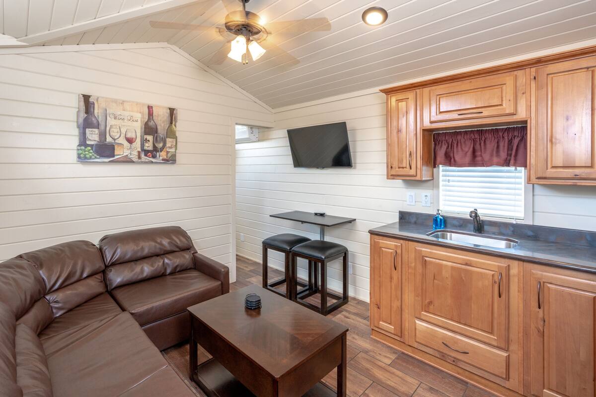 A compact kitchen with wooden cabinets, a built-in TV, and a dining area with a brown leather sofa and a small table.