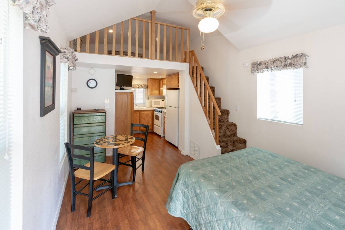 A compact room with a lofted kitchen area, a dining table with chairs, a bed with a quilt, and a staircase.