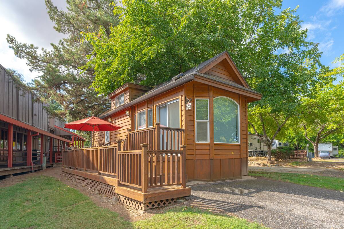 A wooden cabin with a deck and a red umbrella, surrounded by trees and a gravel path.