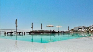 Piscina com espreguiçadeiras e guarda-sóis, cercada por um edifício branco sob um céu azul límpido.